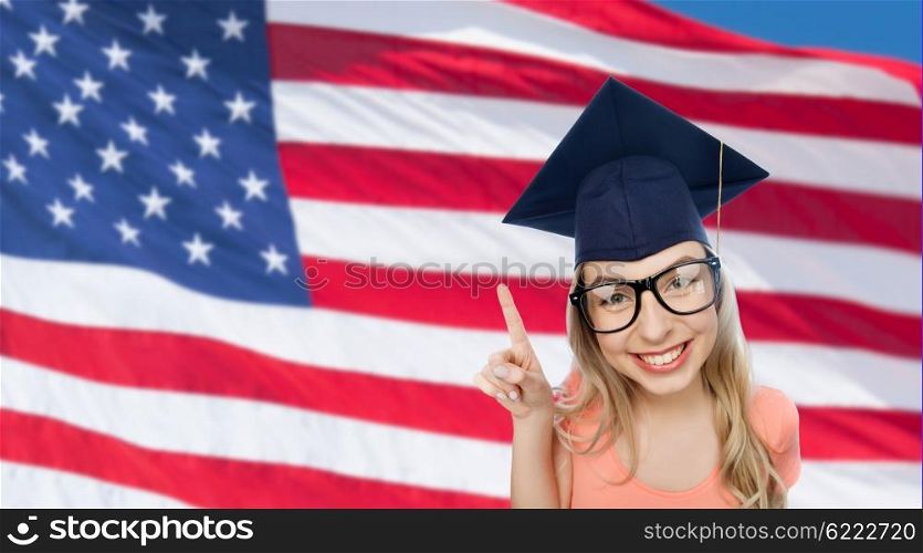 people, graduation and national education concept - smiling young student woman in mortarboard and eyeglasses pointing finger up over american flag background