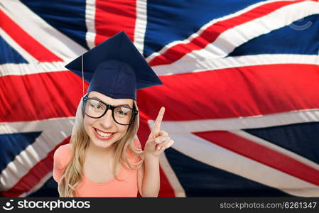 people, graduation and national education concept - smiling young student woman in mortarboard and eyeglasses pointing finger up over english flag
