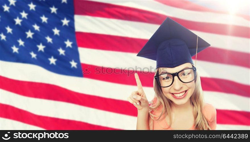 people, graduation and national education concept - smiling young student woman in mortarboard and eyeglasses pointing finger up over american flag background. smiling young student woman in mortarboard