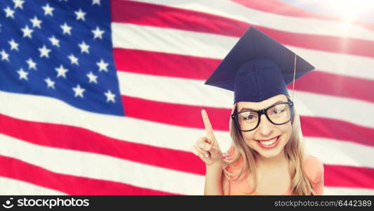 people, graduation and national education concept - smiling young student woman in mortarboard and eyeglasses pointing finger up over american flag background. smiling young student woman in mortarboard