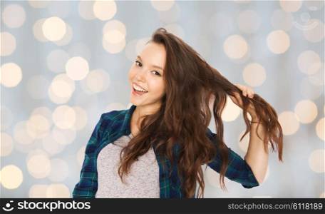 people, hair care, style and teens concept - happy smiling pretty teenage girl holding strand of her hair over holidays lights background