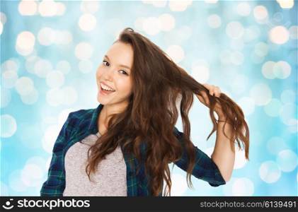 people, hair care, style and teens concept - happy smiling pretty teenage girl holding strand of her hair over blue holidays lights background