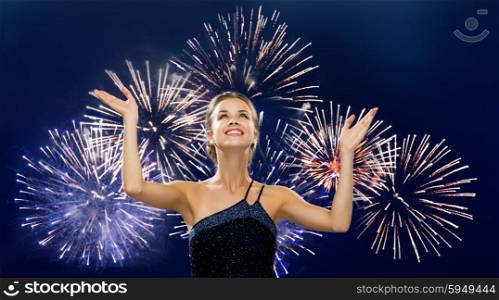 people, happiness, holidays and glamour concept - smiling woman raising hands and looking up over firework on dark blue background