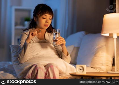 people, health and bedtime concept - stressed asian american woman having headache and taking medicine or sleeping pill with water in bed at night. stressed asian woman taking medicine at night