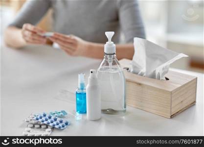 people, health and fever concept - close up of medicines and sanitizers on table over sick woman with thermometer at home. medicines and sick woman with thermometer at home