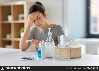 people, health and fever concept - medicines and sanitizers on table over sick woman with thermometer at home. medicines and sick woman with thermometer at home