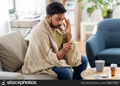 people, health and fever concept - sick indian man in blanket measuring temperature by thermometer at home. sick man measuring temperature by thermometer