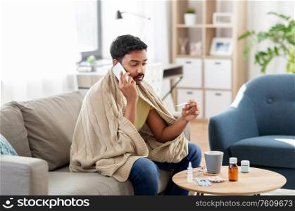 people, health and fever concept - sick indian man in blanket with thermometer calling on smartphone at home. sick man with thermometer calling on smartphone