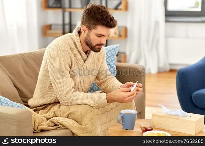 people, health and fever concept - sick man in blanket measuring temperature by thermometer at home. sick man measuring temperature by thermometer