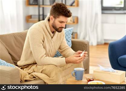 people, health and fever concept - sick man in blanket measuring temperature by thermometer at home. sick man measuring temperature by thermometer