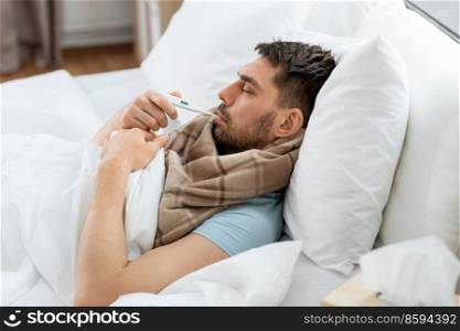 people, health and fever concept - sick man in scarf measuring temperature by thermometer lying in bed at home. sick man measuring temperature by thermometer