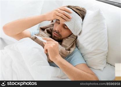 people, health and fever concept - sick man with cold compress on his forehead measuring temperature by thermometer lying in bed at home. sick man measuring temperature by thermometer