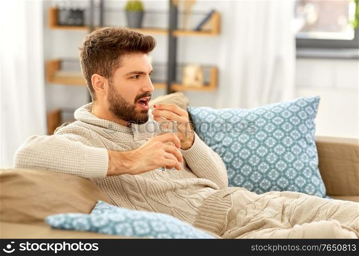 people, health and medicine concept - sad sick man in blanket with glass of water taking pill at home. sick man with glass of water and medicine at home