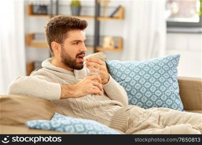 people, health and medicine concept - sad sick man in blanket with glass of water taking pill at home. sick man with glass of water and medicine at home