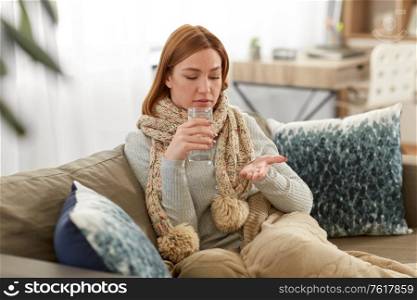 people, health and treatment concept - sad sick young woman in blanket taking medicine with water at home. sick woman taking medicine with water at home