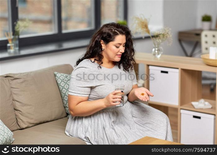 people, health and treatment concept - young woman taking cod liver oil pills with water at home. woman with cod liver oil pills and water at home