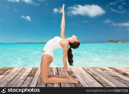 people, health and wellness concept - woman in cotton underwear doing yoga exercise on wooden floor over sea and blue sky background