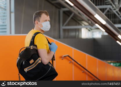 People, health care and coronavirus outbreak concept. Young man wears medical mask and gloves to prevent from pneumonia outbreak, carries backpack on shoulder, concentrated aside, going to travel