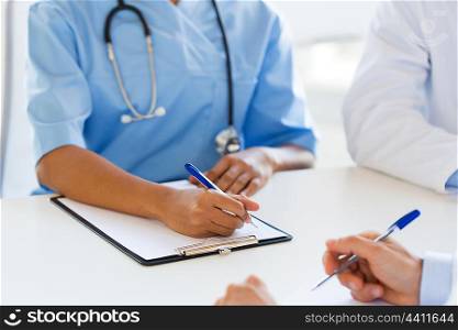 people, health care and medicine concept - close up of african american doctor hands with clipboard and pen writing at hospital