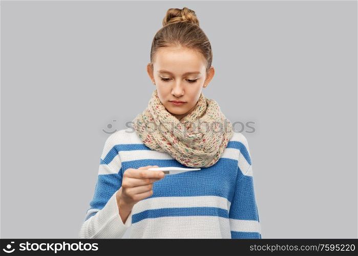 people, health problem and fever concept - sad sick teenage girl in scarf measuring temperature by thermometer over grey background. sick teenage girl in scarf measuring temperature
