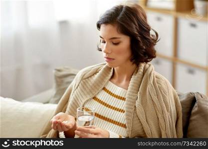 people, health problem and treatment concept - sad sick young woman in blanket taking medicine pills with water at home. sick woman taking medicine with water at home