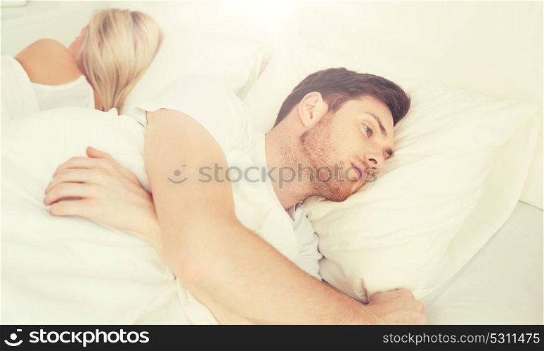 people, health, sleep disorder concept - couple lying back to back in bed at home and young man suffering from insomnia. young man suffering from insomnia