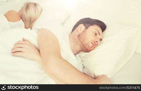 people, health, sleep disorder concept - couple lying back to back in bed at home and young man suffering from insomnia. young man suffering from insomnia