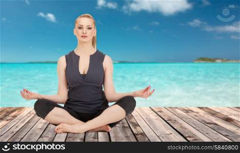 people, health, wellness and meditation concept - happy young woman meditating in yoga lotus pose on wooden floor over sea and blue sky background