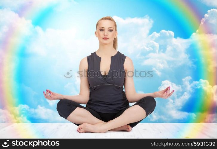 people, health, wellness and meditation concept - happy young woman meditating in yoga lotus pose on wooden floor over white clouds and rainbow on blue sky background