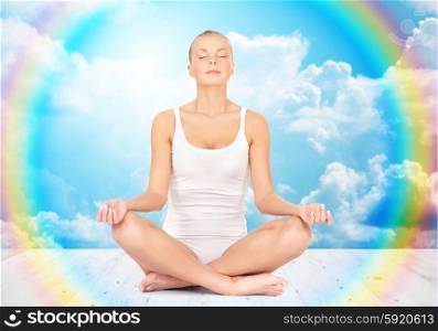 people, health, wellness and meditation concept - woman in underwear meditating in yoga lotus pose on wooden floor over white clouds and rainbow on blue sky background