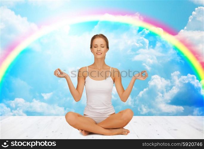 people, health, wellness and meditation concept - woman in underwear meditating in yoga lotus pose on wooden floor over white clouds and rainbow on blue sky background