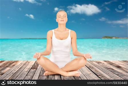 people, health, wellness and meditation concept - woman in underwear meditating in yoga lotus pose on wooden floor over sea and blue sky background