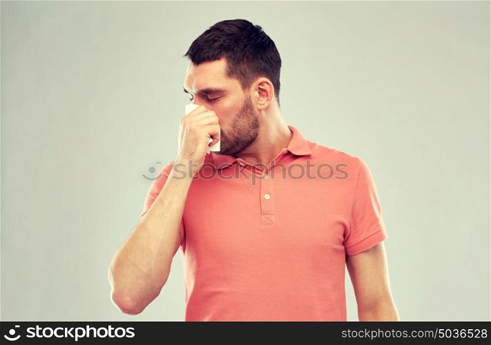 people, healthcare, rhinitis, cold and allergy concept - sick man with paper napkin blowing nose over gray background. sick man with paper napkin blowing nose