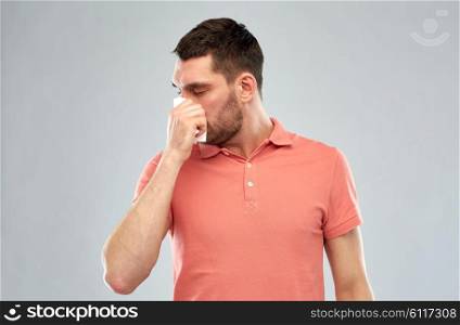 people, healthcare, rhinitis, cold and allergy concept - sick man with paper napkin blowing nose over gray background