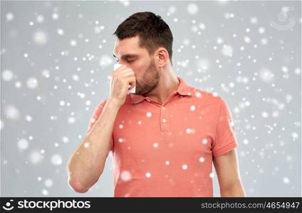 people, healthcare, rhinitis, winter and cold concept - sick man with paper wipe blowing nose over snow on gray background