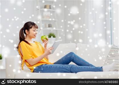 people, healthy eating, education, technology and concept - happy young asian woman sitting on sofa with tablet pc computer and green apple at home over snow