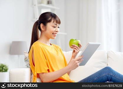 people, healthy eating, education, technology and concept - happy young asian woman sitting on sofa with tablet pc computer and green apple at home