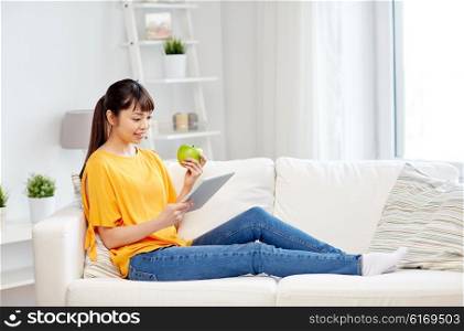 people, healthy eating, education, technology and concept - happy young asian woman sitting on sofa with tablet pc computer and green apple at home