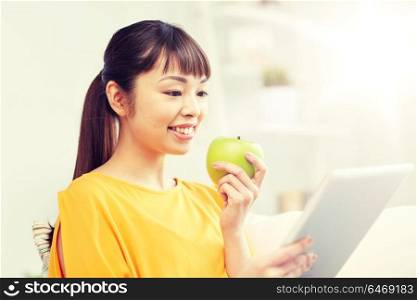 people, healthy eating, education, technology and concept - happy young asian woman sitting on sofa with tablet pc computer and green apple at home. happy asian woman with tablet pc and apple at home