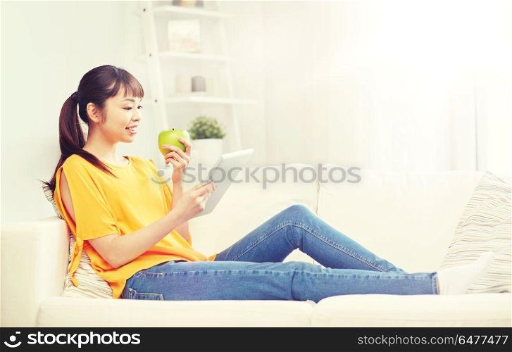 people, healthy eating, education, technology and concept - happy young asian woman sitting on sofa with tablet pc computer and green apple at home. happy asian woman with tablet pc and apple at home. happy asian woman with tablet pc and apple at home
