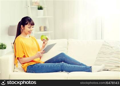 people, healthy eating, education, technology and concept - happy young asian woman sitting on sofa with tablet pc computer and green apple at home. happy asian woman with tablet pc and apple at home. happy asian woman with tablet pc and apple at home