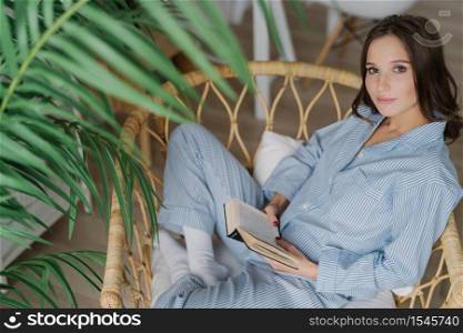 People, hobby and comfort concept. Top view of brunette woman sits in lotus pose on wicker chair, reads interesting book, enjoys domestic atmosphere, looks with relaxed expression at camera.