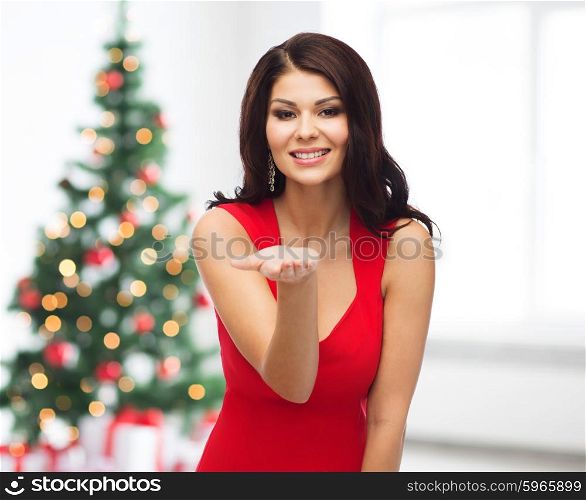 people, holidays and advertisement concept - beautiful sexy woman in red dress showing something on empty hand over room with christmas tree and gifts background