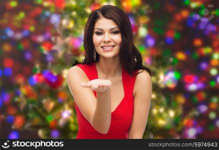 people, holidays and advertisement concept - beautiful sexy woman in red dress showing something on empty hand over christmas party lights background