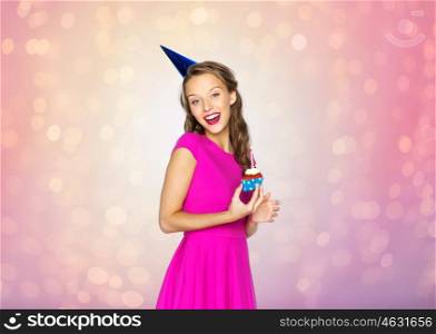 people, holidays and celebration concept - happy young woman or teen girl in pink dress and party cap with birthday cupcake over rose quartz and serenity lights background