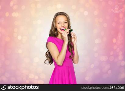 people, holidays and celebration concept - happy young woman or teen girl in pink dress and party horn or blower over rose quartz and serenity lights background