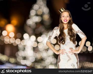people, holidays and celebration concept - happy young woman or teen girl in party dress and princess crown over christmas tree lights background. happy woman in crown over christmas tree lights