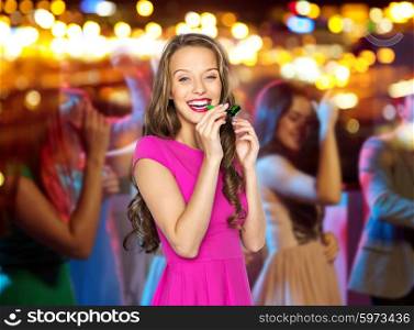people, holidays and celebration concept - happy young woman or teen girl in pink dress blowing to party horn at night club over crowd and lights background