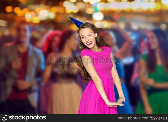 people, holidays and celebration concept - happy young woman or teen girl in pink dress and party cap at night club party over crowd and lights background