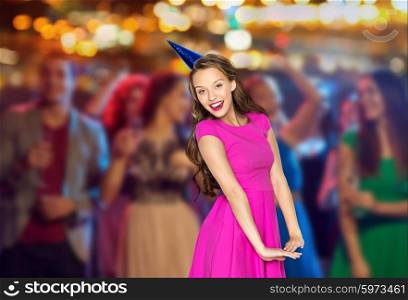 people, holidays and celebration concept - happy young woman or teen girl in pink dress and party cap at night club party over crowd and lights background